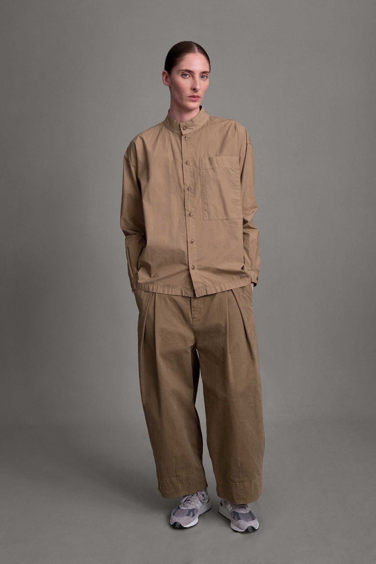Toogood The Physician Shirt. Model on grey backdrop wears light brown button-down shirt with brown pants and sneakers.