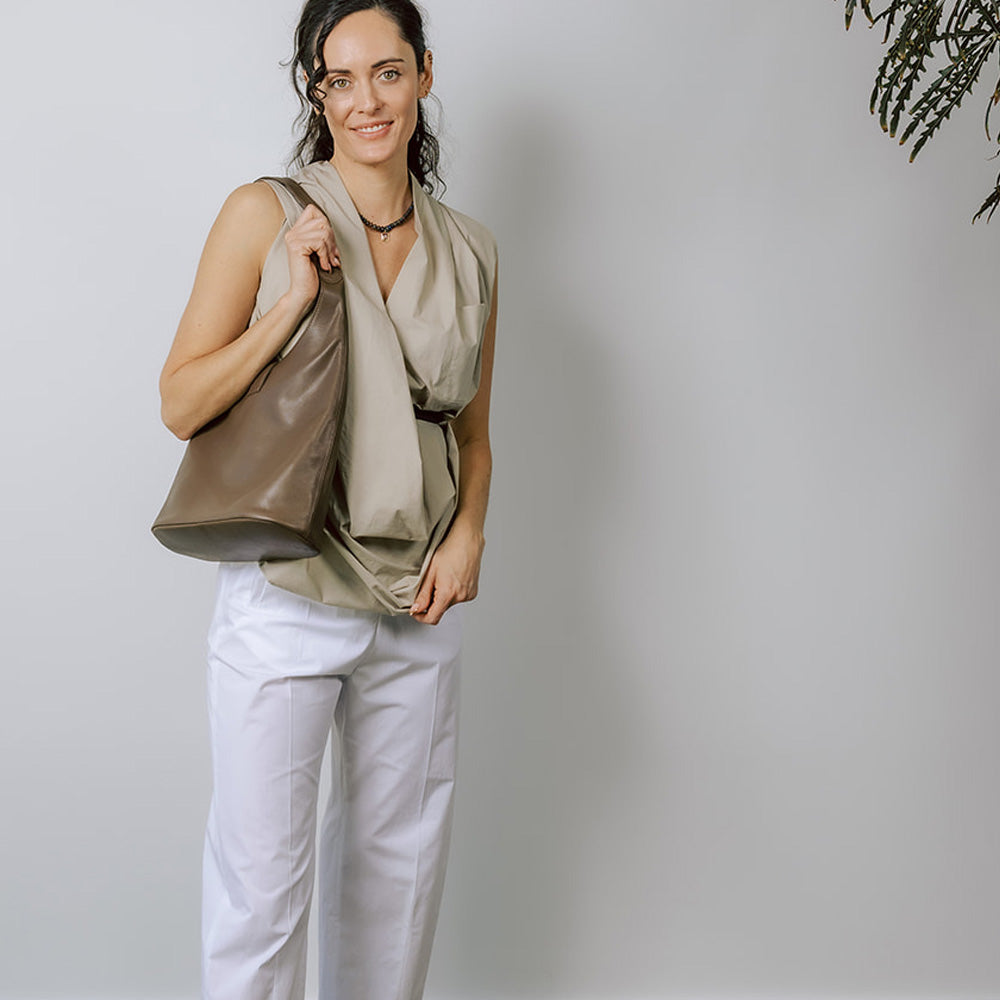 Model on grey backdrop wears brown leather ARE STUDIO Sophia handbag with beige pleated top and white pants