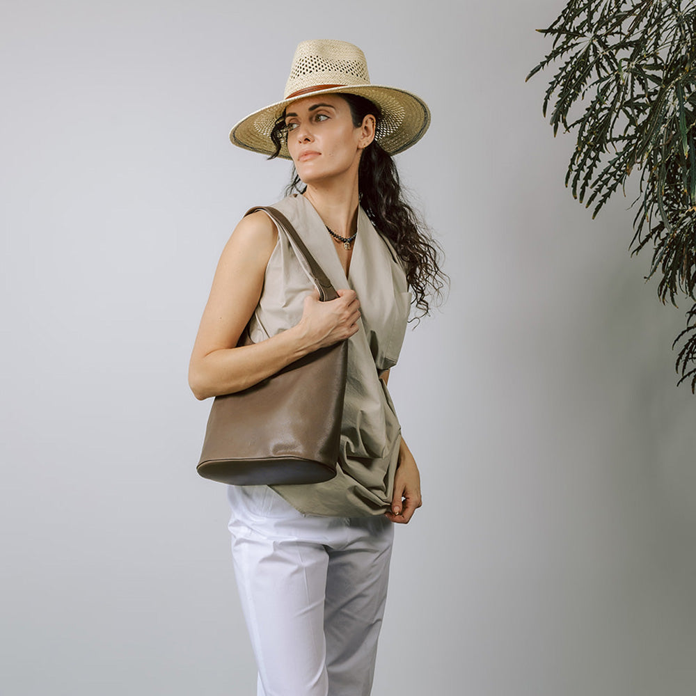 Model on grey backdrop wears brown leather ARE STUDIO Sophia handbag with beige pleated top and hat