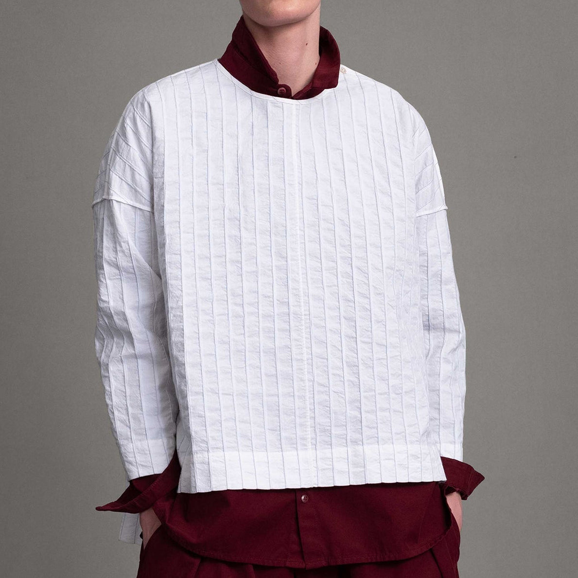 Alt model on grey backdrop wears white Archivist top over maroon button-down shirt and pants