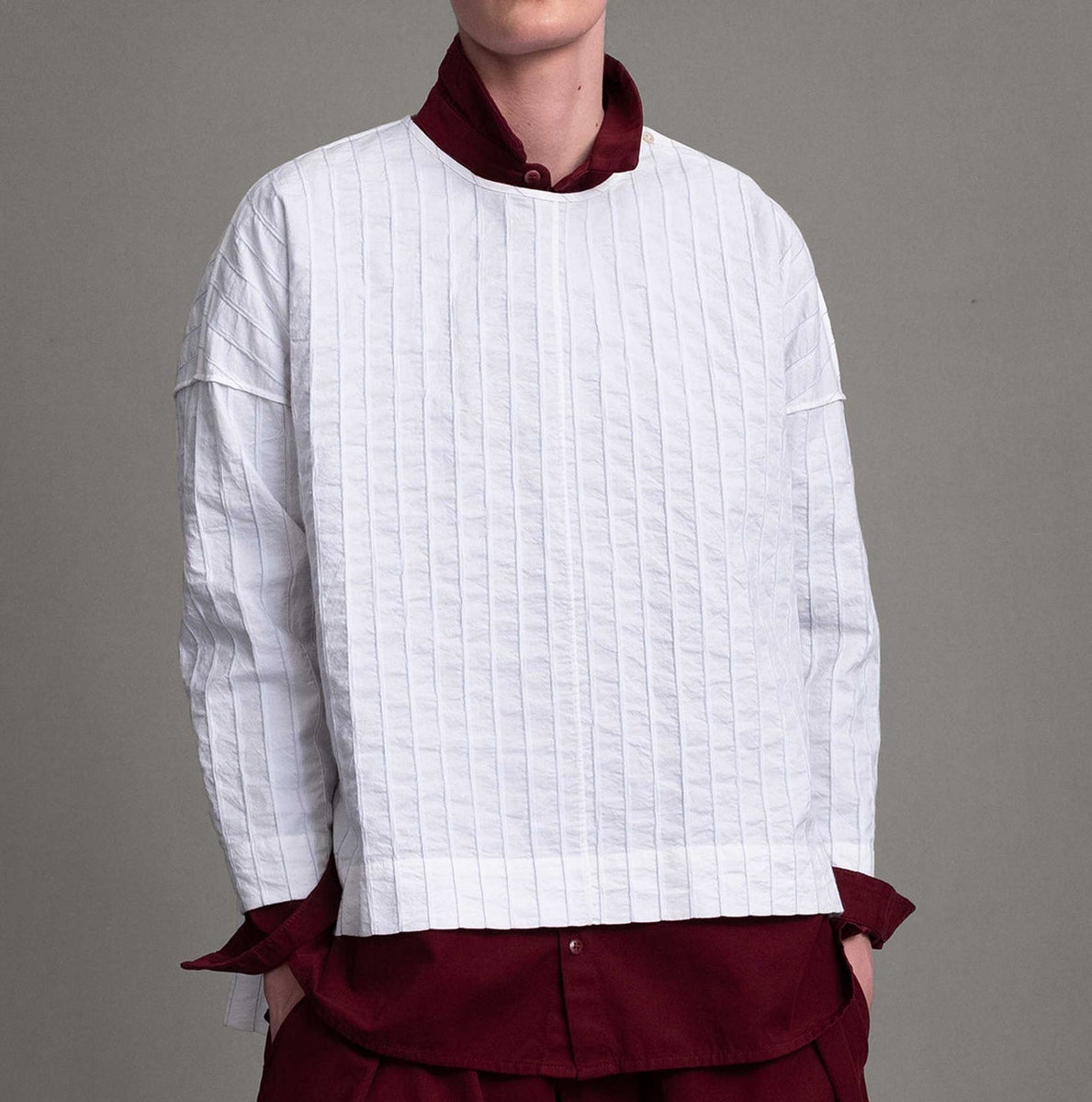 Alt model on grey backdrop wears white Archivist top over maroon button-down shirt and pants