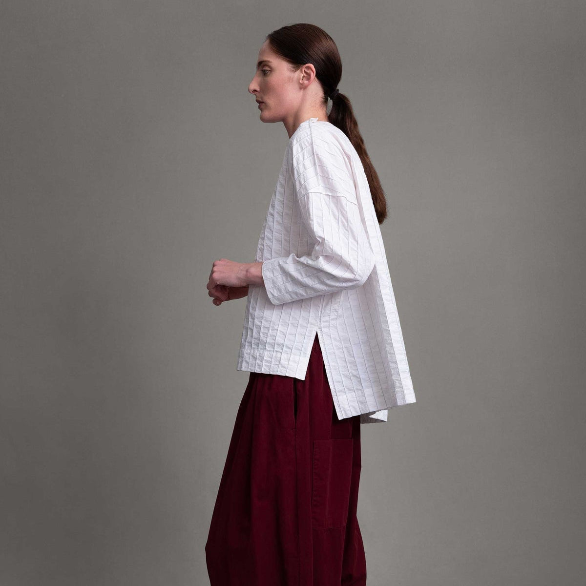 Side view of model on grey backdrop wearing white Archivist top with maroon pants