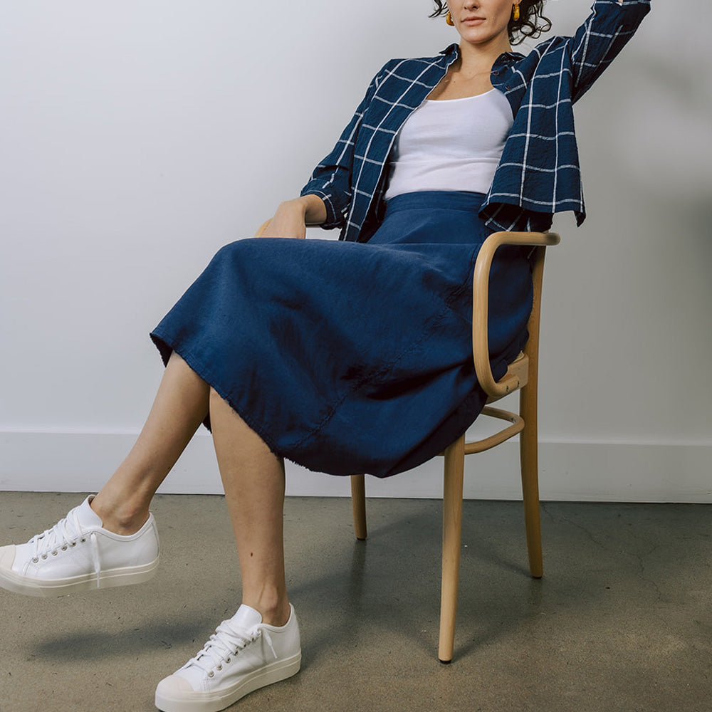 Model sits in chair and wears H+ HANNOH WESSEL blue Jocelyne midi skirt with blue checked shirt and white sneakers