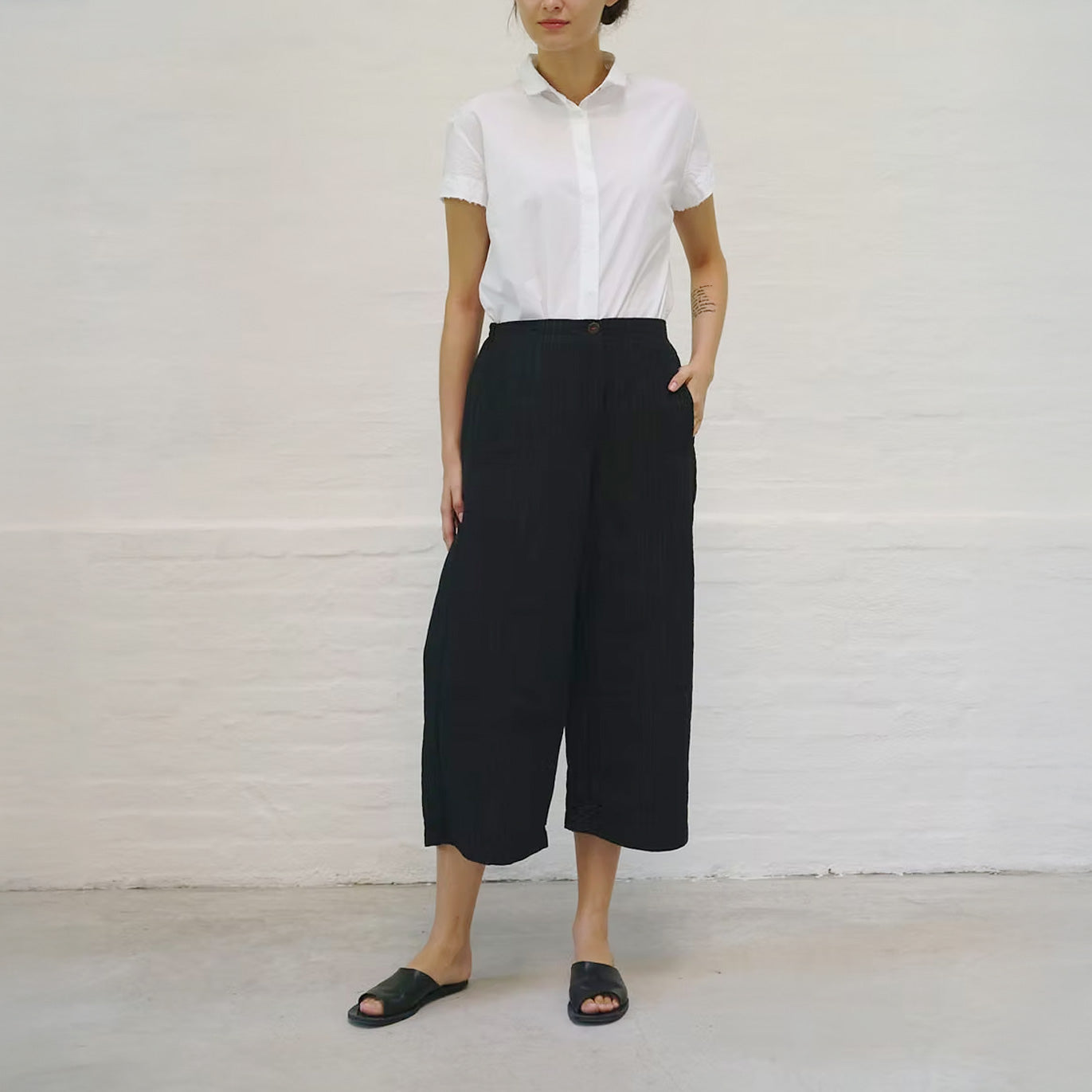 Model on white backdrop wears H+ HANNOH WESSEL black wide-leg cropped Poppy pants and white shirt
