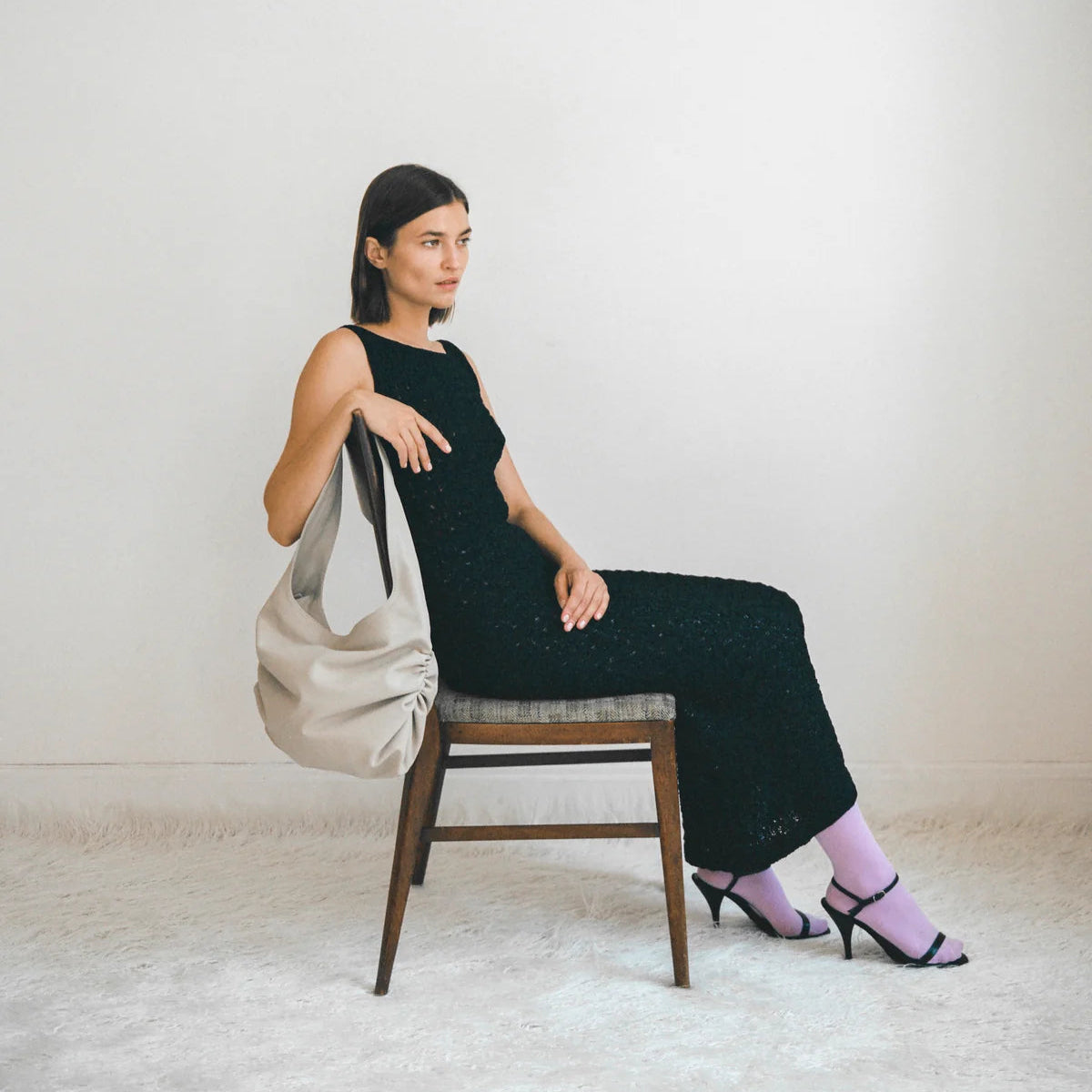 Model sits in chair in white studio, wears black dress with grey Stella Bag slung on back of chair.