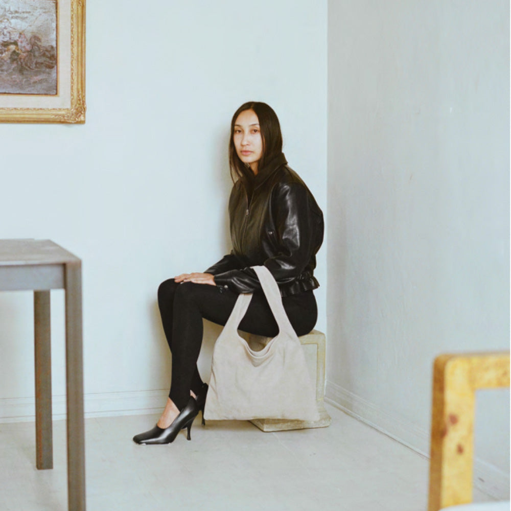 Model in black outfit sits in corner with white Stella bag over her arm.
