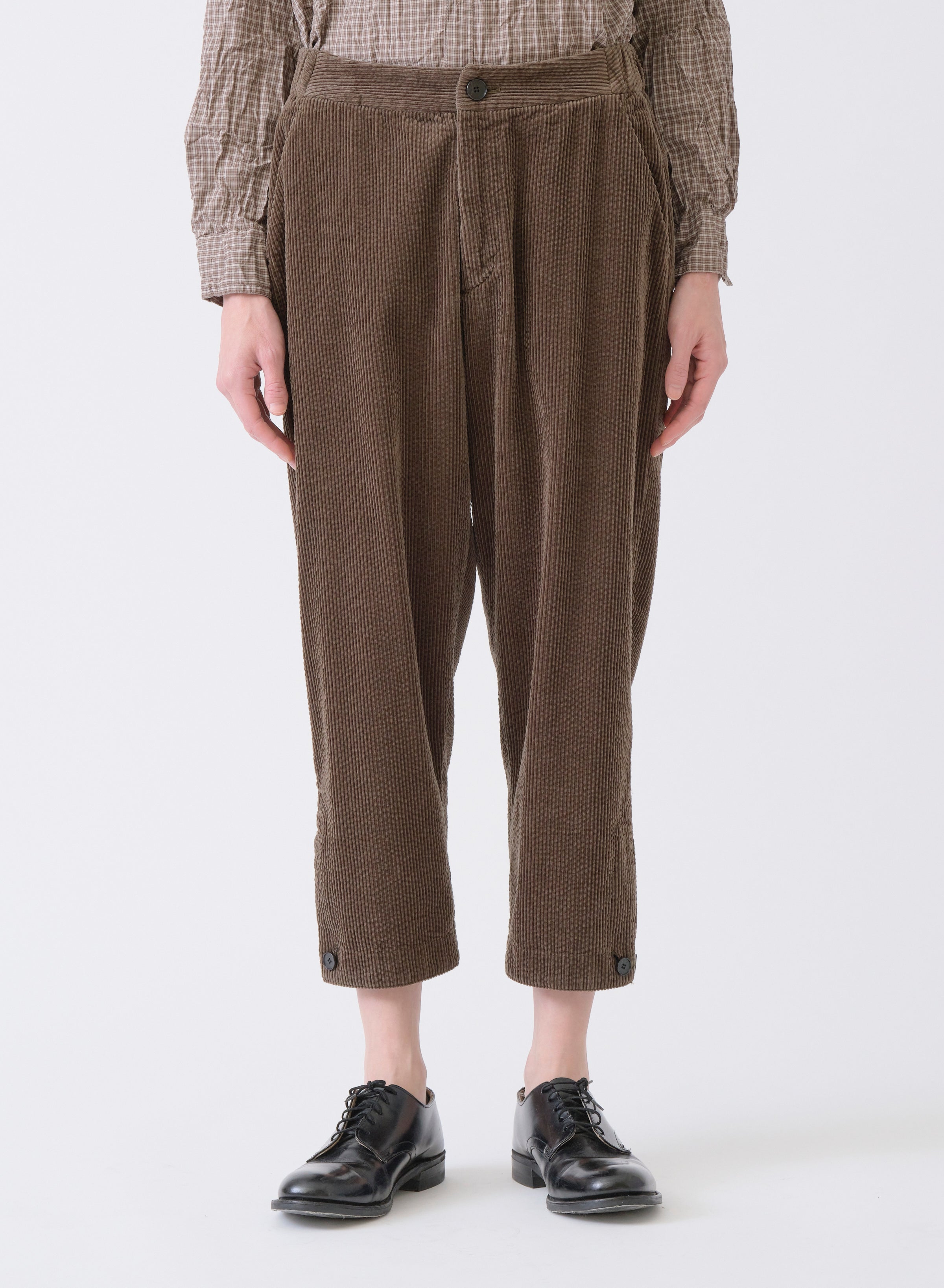 Close-up of model on grey backdrop wearing PAS DE CALAIS brown cropped corduroy tapered pants and black shoes