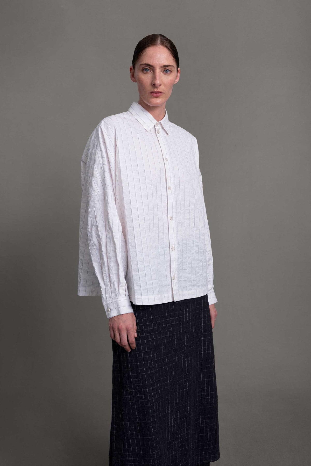 Model on grey backdrop wears boxy white Toogood The Farrier Shirt with buttons, and long dark skirt, closer view.