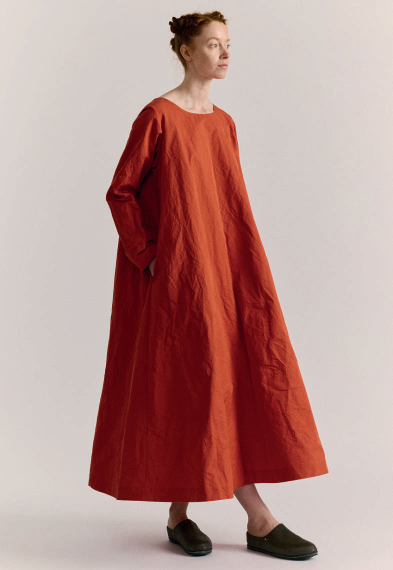 Model on grey backdrop wears Casey Casey dress in rust orange with long sleeves, oversized cut