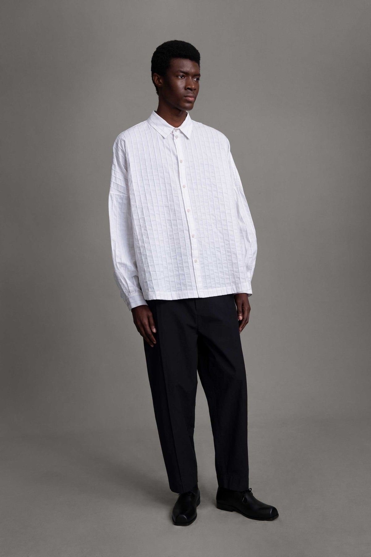 Model on grey backdrop wears white Toogood The Farrier button-down Shirt with black pants.