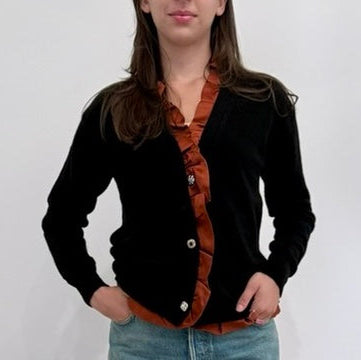 Model on grey backdrop with hands at hips wears Leur Logette black cashmere stretch cardigan with rust ruffle details, and jeans
