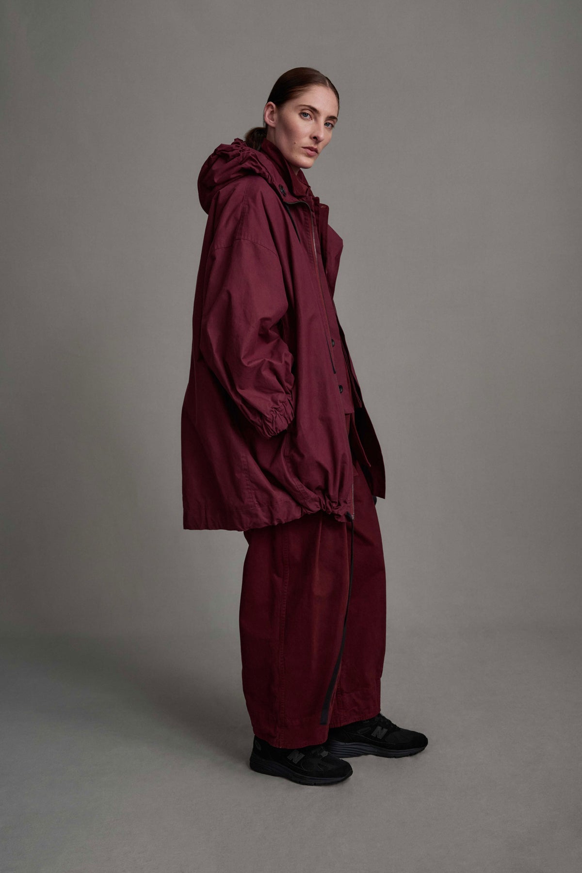 Side view of model on grey backdrop in oversized Toogood Explorer Coat in maroon, with matching color pants