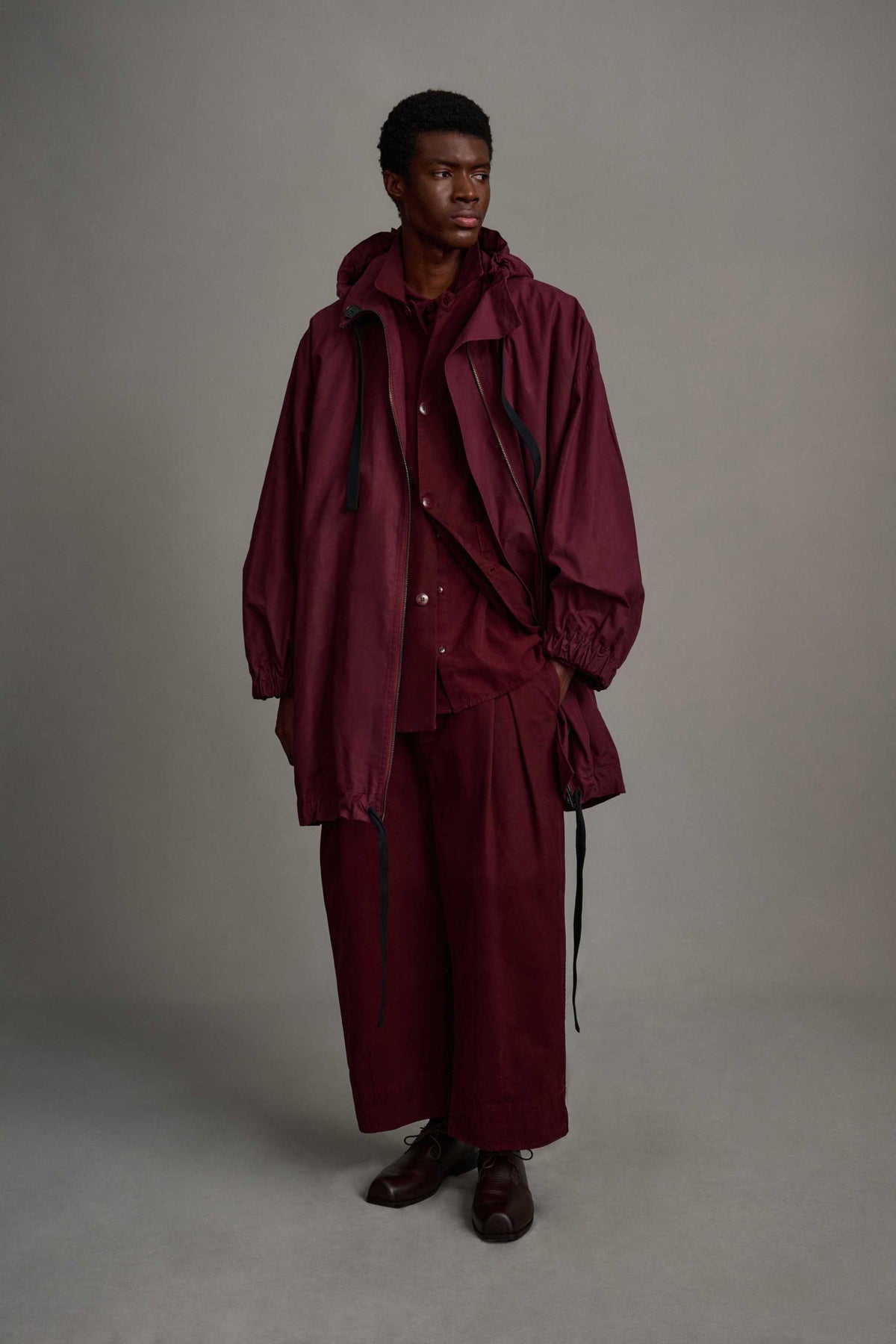 Male model on grey backdrop wears Toogood Explorer Coat in maroon and matching color pants