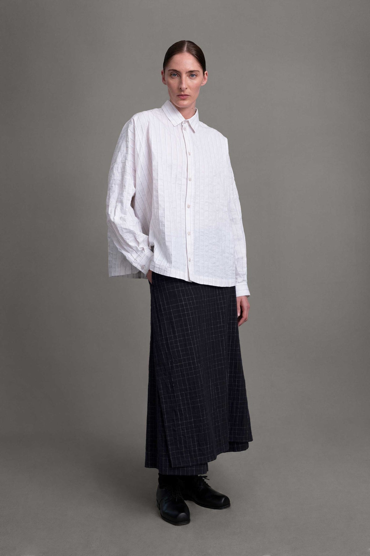 Model on grey backdrop wears boxy white Toogood The Farrier Shirt with buttons, and long dark skirt