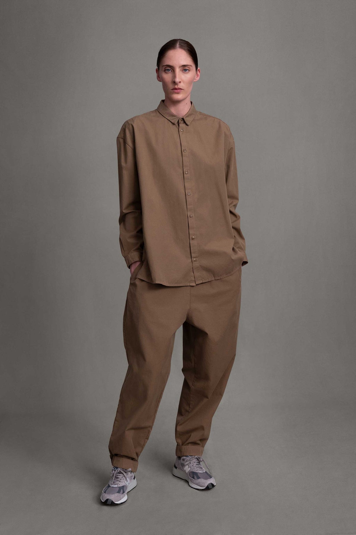 Model on grey backdrop wears Toogood The Gymnast Trouser in light brown, with matching button-down and sneakers.
