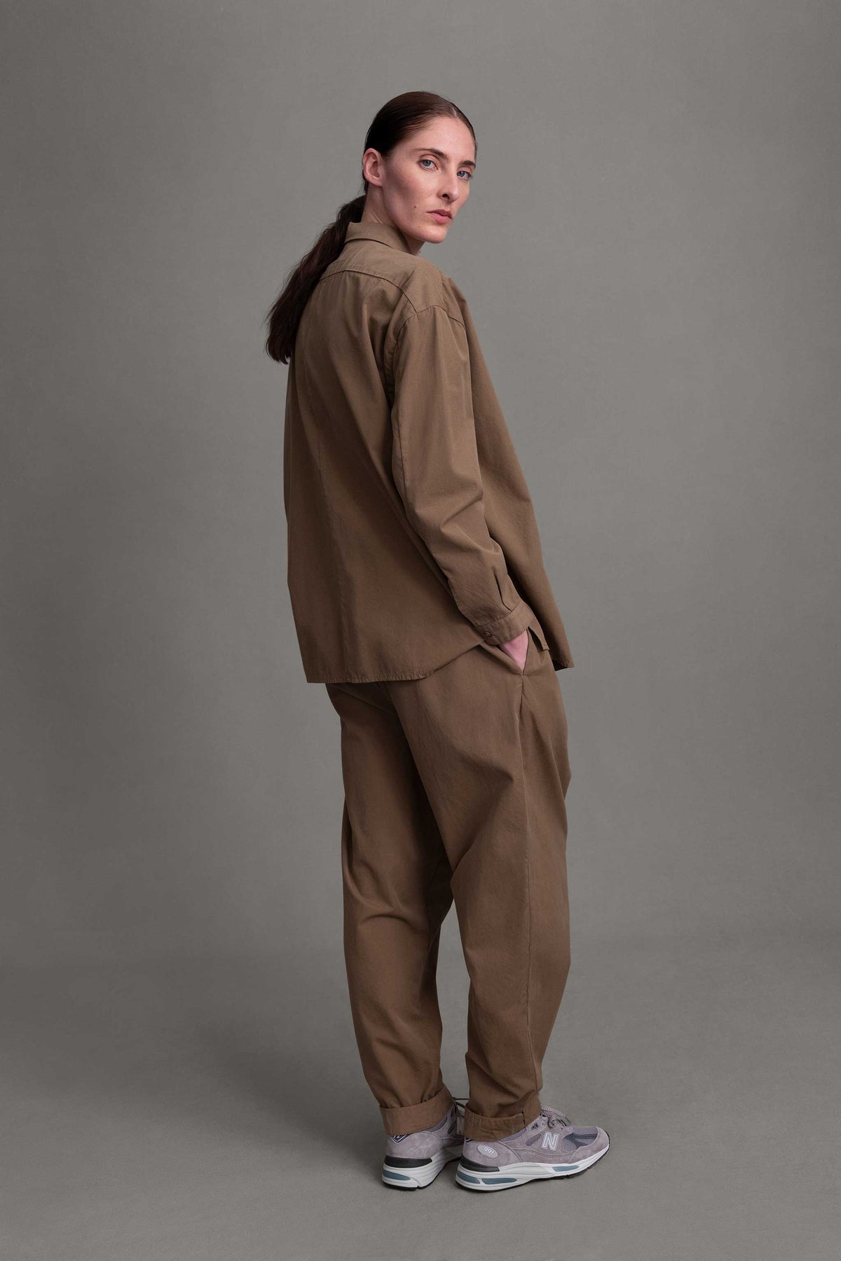 Back view of model on grey backdrop in Toogood light brown Trouses, with matching button-down and sneakers.
