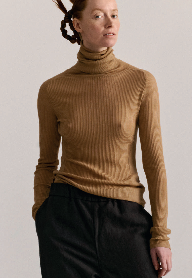 Model on grey backdrop wear Casey Casey merino wool rib turtleneck in light brown with black pants