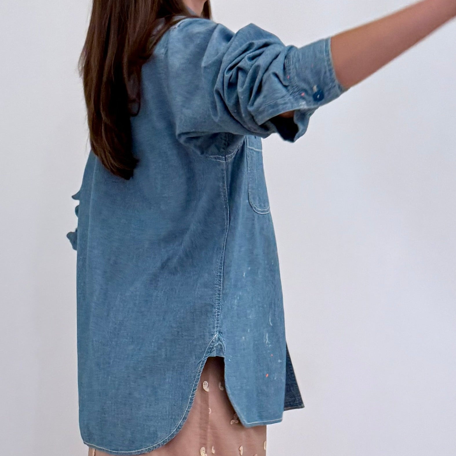Side view of Model on grey backdrop wearing CHIMALA blue chambray work shirt over pink skirt