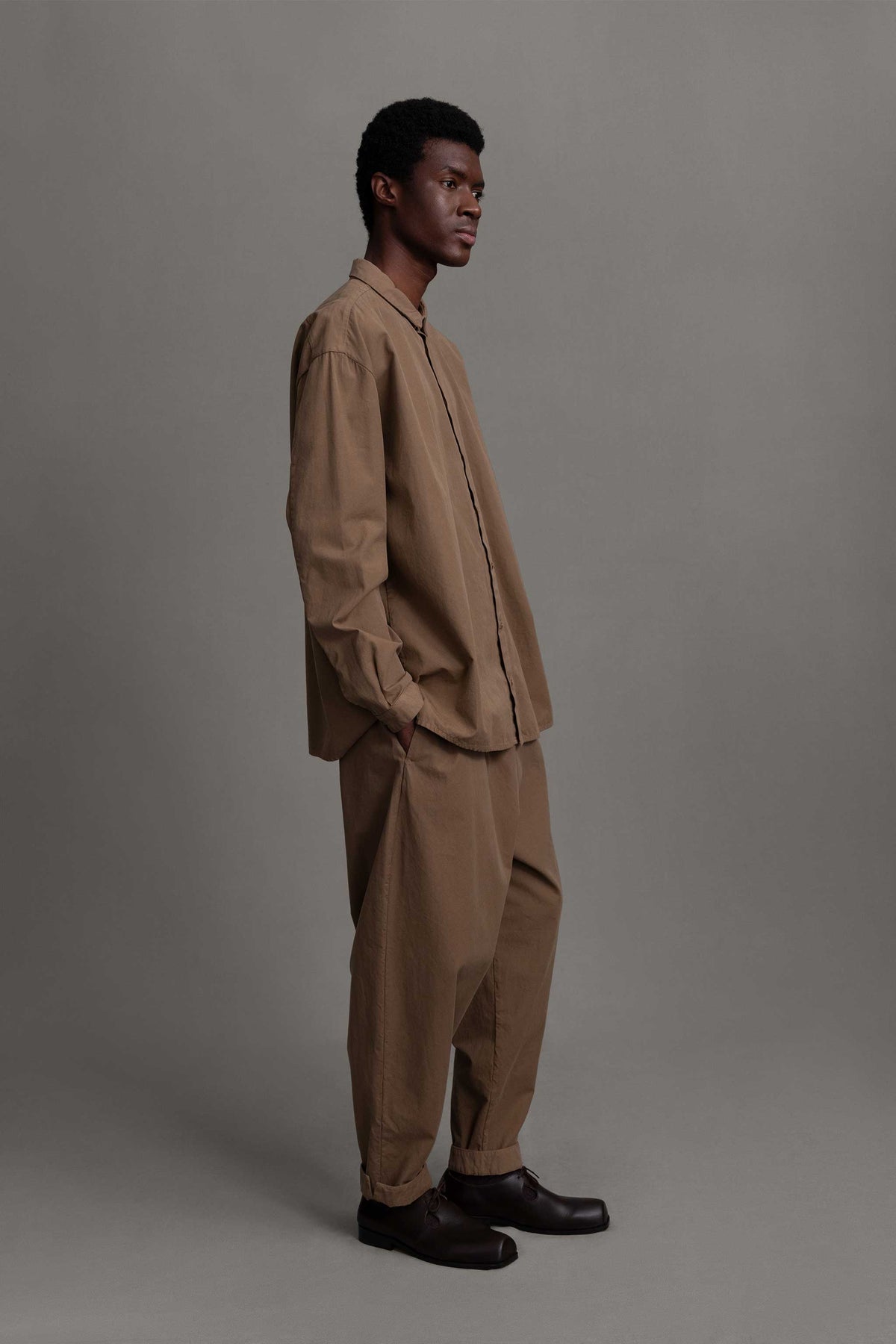 Side view of male model on grey backdrop in Toogood light brown Trouses, with matching button-down and brown shoes
