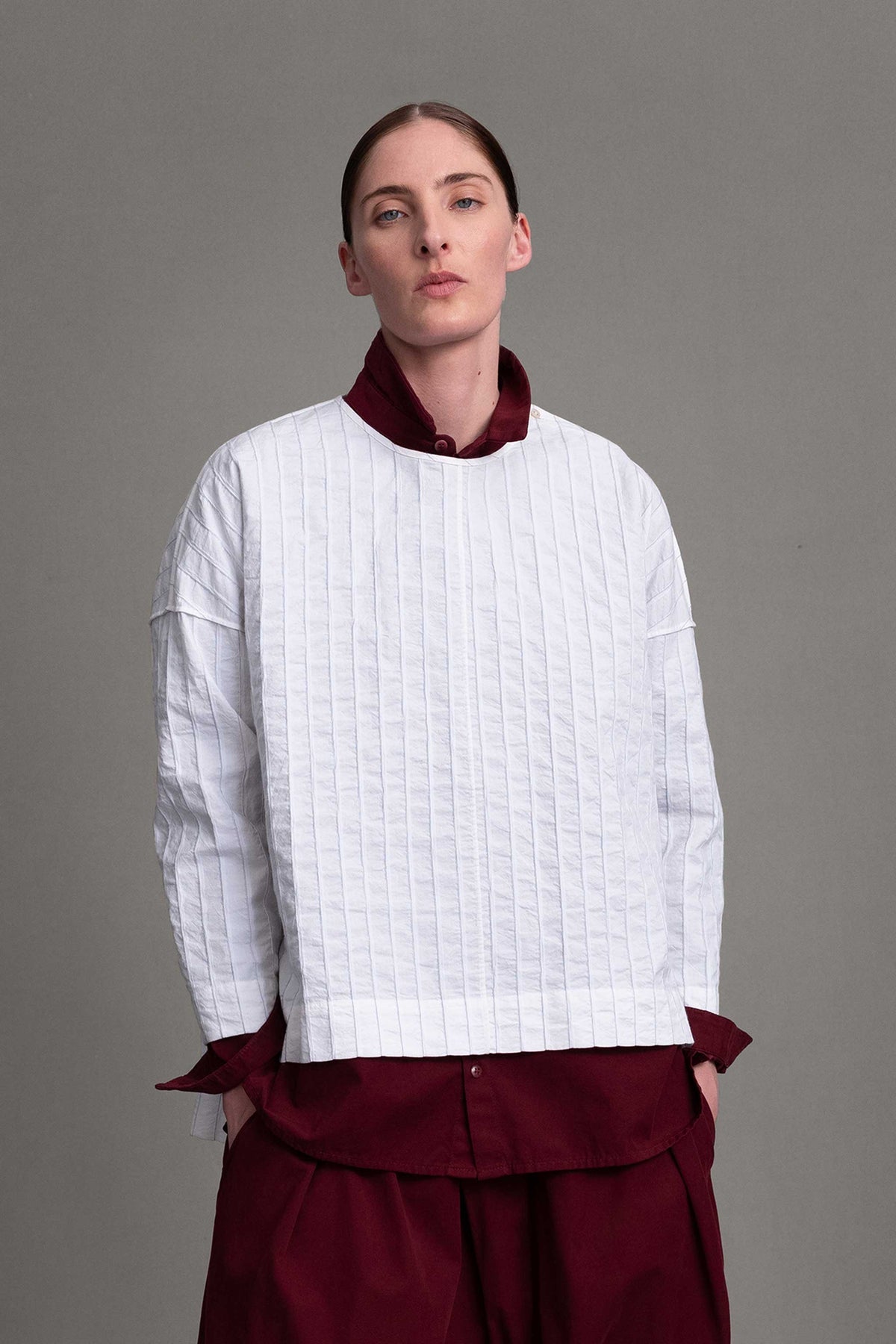 Alt model on grey backdrop wears white Archivist top over maroon button-down shirt and pants