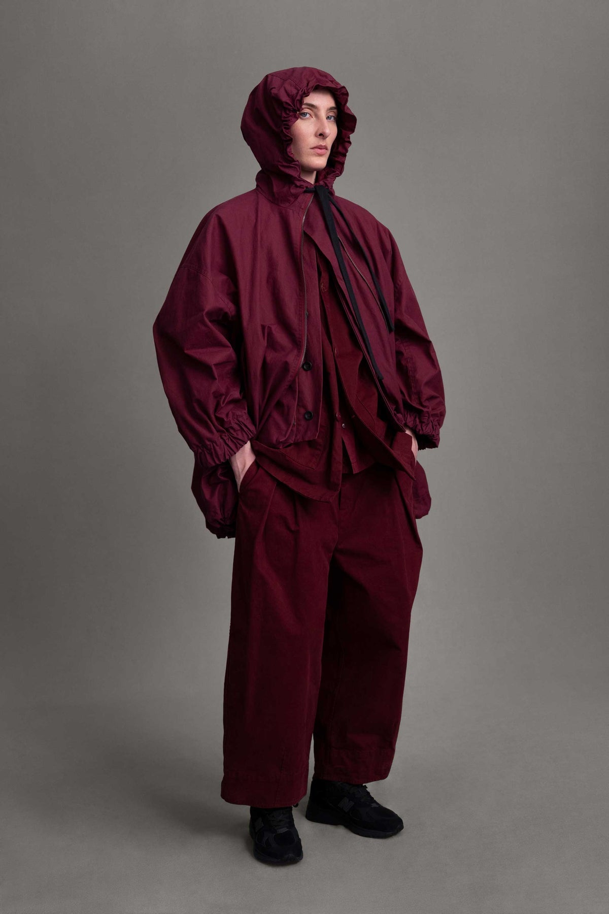 Model on grey backdrop wears oversized Toogood Explorer Coat in maroon with hood up and matching color pants