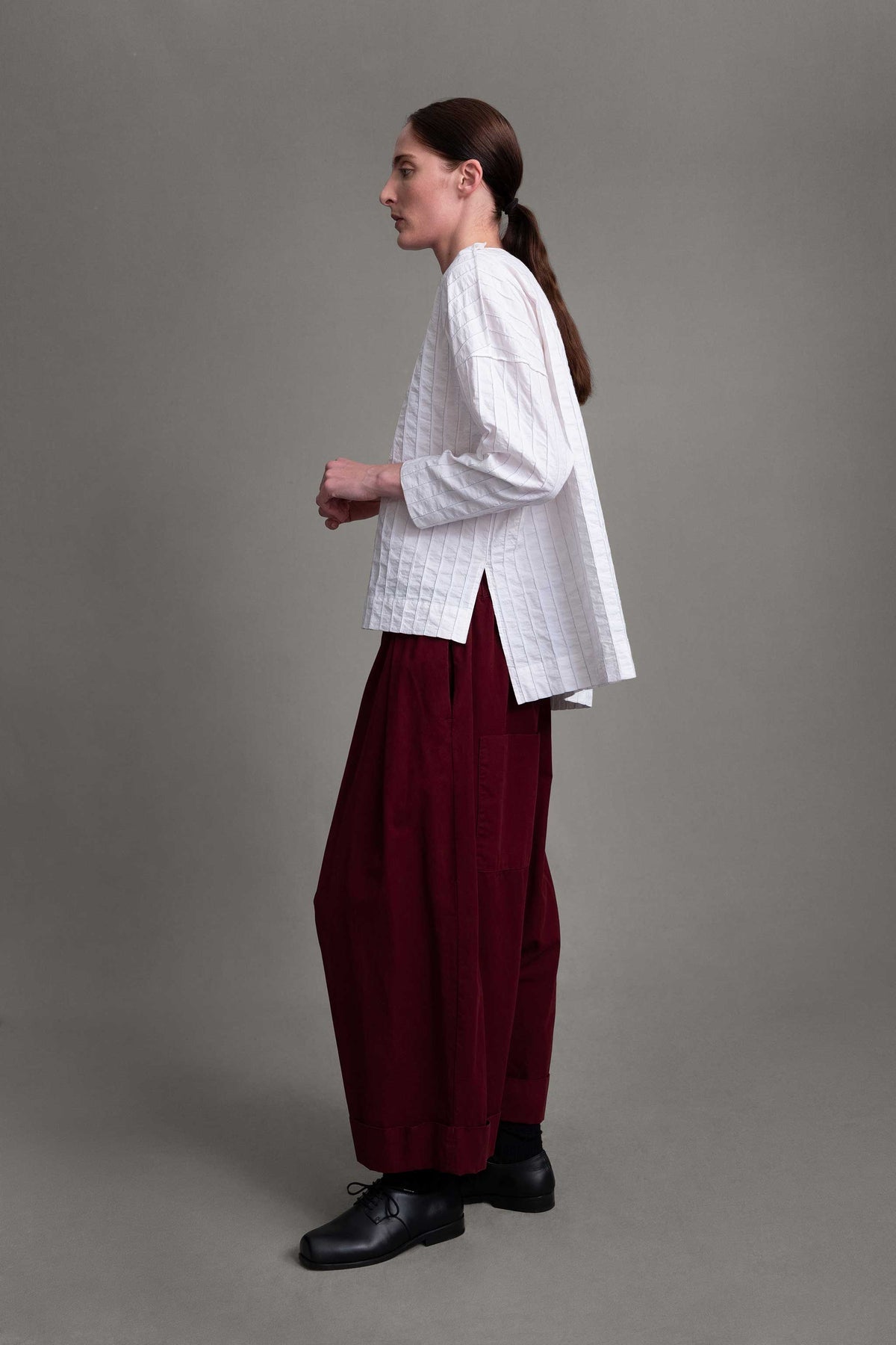 Side view of model on grey backdrop wearing white Archivist top with maroon pants