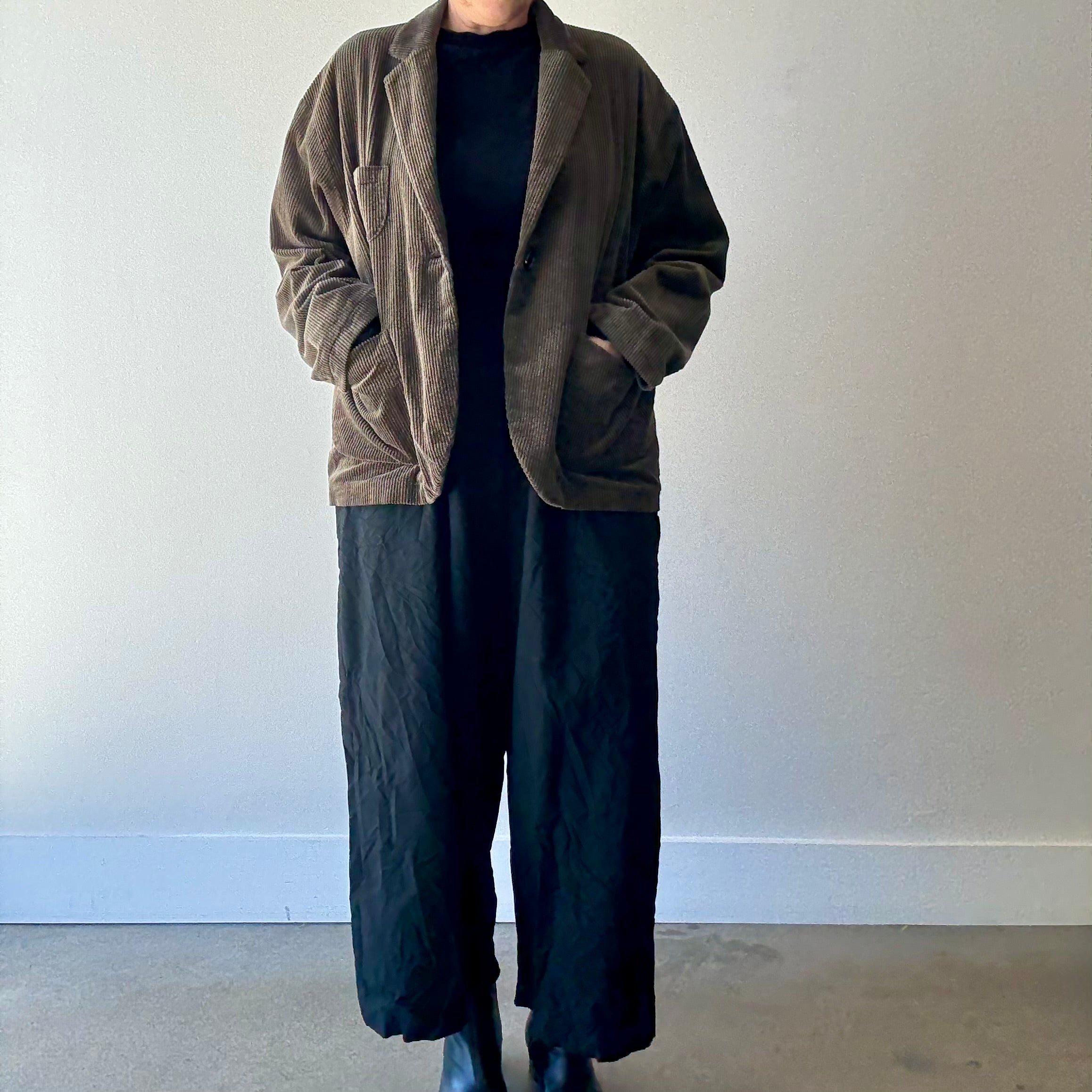Model in studio from neck down wears PAS DE CALAIS brown cotton corduroy jacket with black shirt and pants