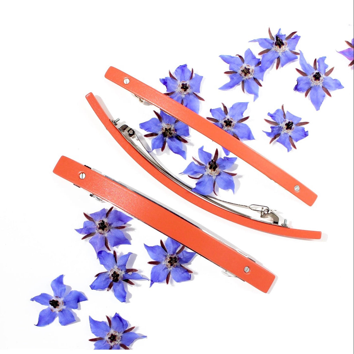 Three SYLVAIN LE HEN barrettes in orange surrounded by purple flowers, on white backdrop