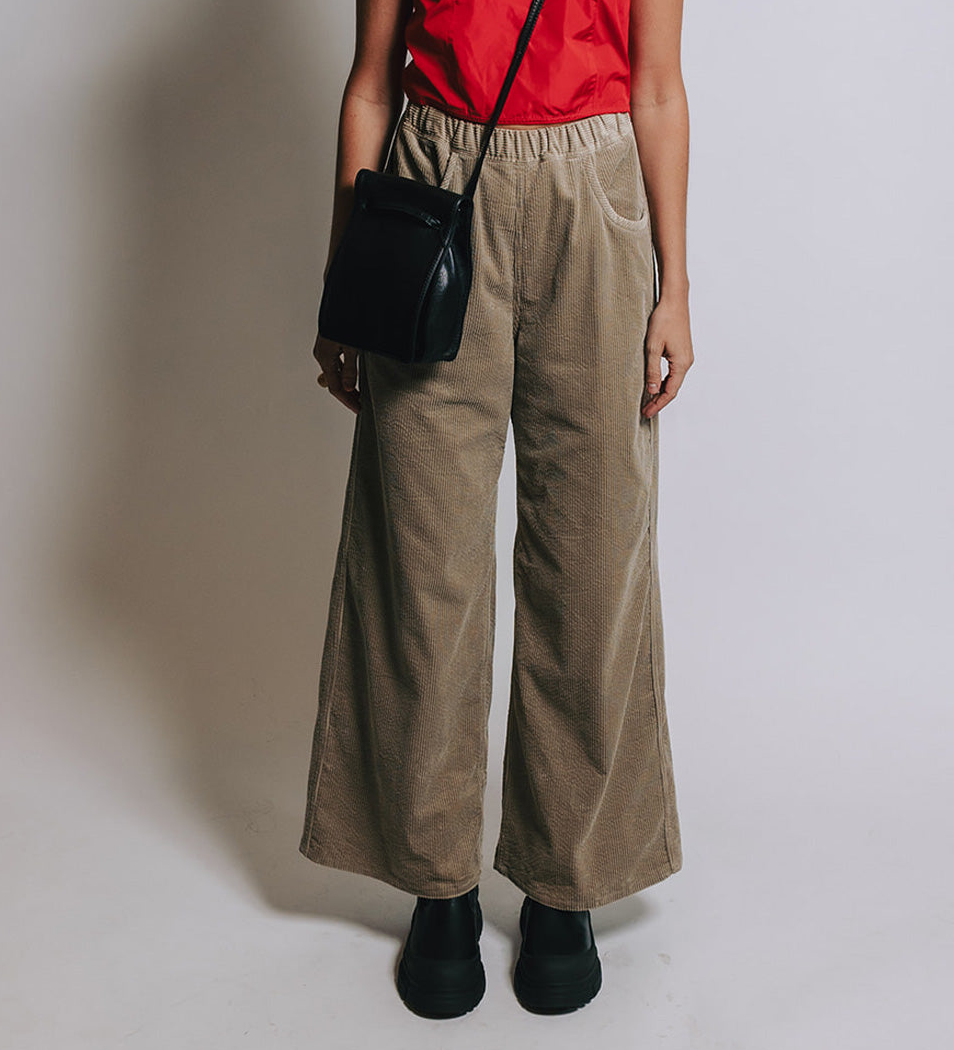Model on grey backdrop wearing SOFIE D'HOORE brown corduroy Pistis Pants with red shirt and black bag