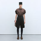 Cordera Tailoring Bermuda Shorts. Model wears black shorts with brown button-down and fashion hat.