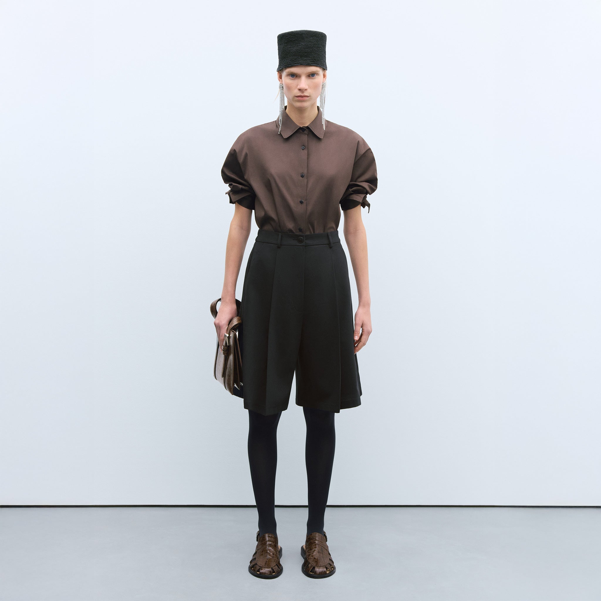 Cordera Tailoring Bermuda Shorts. Model wears black shorts with brown button-down and fashion hat.